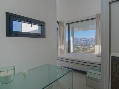 Dormitorio luminoso con cama frente a ventanales de suelo a techo, revelando vistas panorámicas a la montaña y el valle. Se aprecia una consola de cristal y una ventana horizontal con marco oscuro.