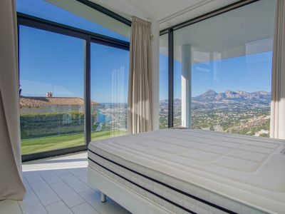 Dormitorio moderno con cama grande y amplios ventanales. Ofrece vistas panorámicas al mar distante, un césped verde y montañas escarpadas bajo un cielo despejado.
