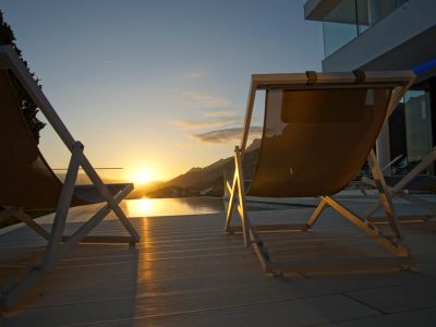 Dos tumbonas modernas sobre tarima clara frente a una piscina infinita que refleja el atardecer dorado sobre las montañas.