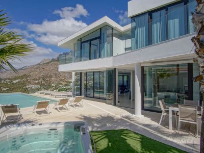 El exterior de la villa moderna blanca presenta una piscina infinita, jacuzzi independiente y una amplia terraza con tumbonas, enmarcado por vistas a la montaña bajo un cielo despejado.