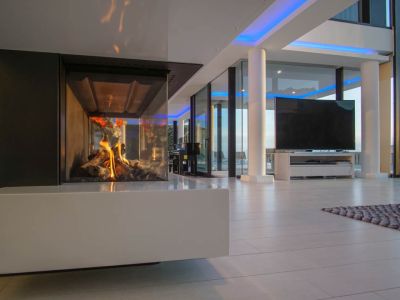 Espacio de salón moderno con chimenea de gas acristalada y base blanca. Grandes ventanales y luz ambiental azul complementan el diseño.
