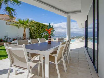 Esta terraza comedor exterior cubierta presenta una mesa blanca moderna con ocho sillas beige sobre baldosas claras. Ofrece amplias vistas a las montañas y al mar, junto a la piscina infinita.