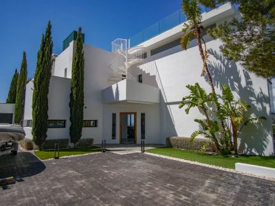 Exterior de la villa moderna con entrada pavimentada, cipreses altos y una escalera de caracol blanca que asciende a la terraza superior con barandilla de cristal.