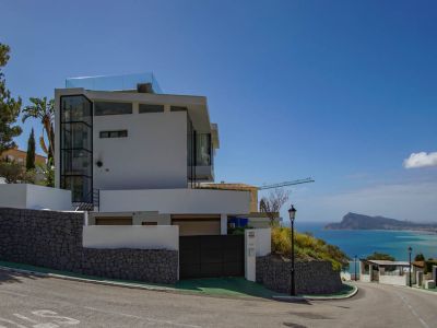 Exterior de villa moderna blanca con ascensor de cristal visible y barandilla de terraza en la azotea. Muros de piedra y portón automático complementan las vistas panorámicas al mar.