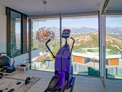 Gimnasio con ventanales de suelo a techo, ofreciendo vistas panorámicas a las montañas y al valle. Equipado con elíptica y pesas, el espacio incluye una barandilla de cristal y un diseño moderno.
