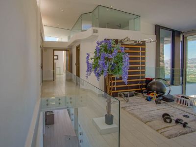 Gimnasio en planta superior con barandales de cristal modernos y suelo claro. Incluye escalera sueca y pesas, ofreciendo vistas panorámicas a través de amplios ventanales de suelo a techo.