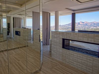 Interior moderno con chimenea de gas transparente y base de guijarros, integrada en una pared texturizada. Paneles espejados reflejan la estancia y las amplias vistas a la montaña y al mar.