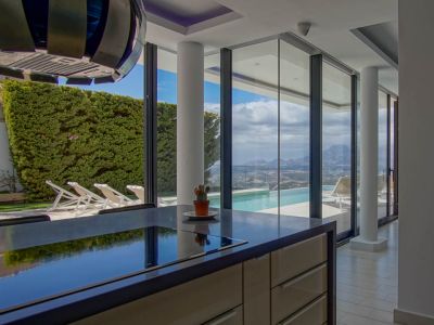 Isla de cocina moderna con encimera oscura y placa integrada, frente a grandes ventanales con vistas a la piscina infinita, el valle y las montañas.
