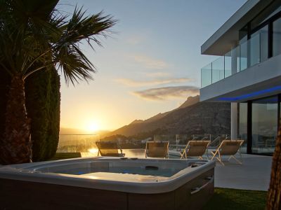 Jacuzzi exterior con acabado de madera, rodeado de césped, frente a una villa moderna con balcones de cristal. Vistas a la montaña y al mar al atardecer.