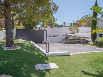 La entrada principal presenta un portón automático oscuro y un área de estacionamiento pavimentada con una embarcación. El césped artificial y un pino maduro complementan el exterior.