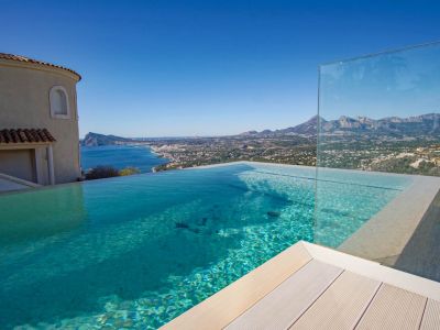 La piscina infinita de agua cristalina y cubierta clara, con barandilla de cristal, ofrece vistas panorámicas al mar, la costa y las montañas bajo un cielo despejado.