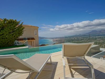 La terraza exterior presenta una piscina infinita con vistas a la costa y montañas lejanas. Dos tumbonas sobre tarima clara, enmarcadas por barandilla de cristal y fachada de villa tradicional.