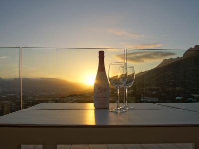 Mesa exterior en terraza con barandilla de cristal, presentando una botella de cava y dos copas. Atardecer sobre montañas y valle, con el sol poniente reflejándose.