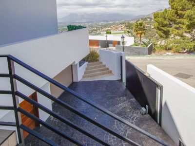 Moderna escalera exterior con barandales negros desciende a un camino de entrada texturizado en gris oscuro, con puerta automática, acceso a garaje y vistas panorámicas a la montaña.