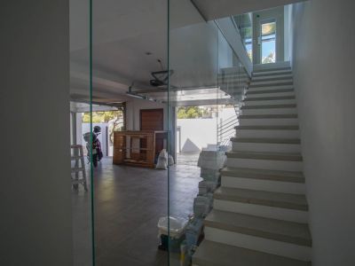 Moderna escalera interior con barandilla de cristal y peldaños claros, adyacente a un garaje con suelo de baldosas y puerta abierta con luz natural.