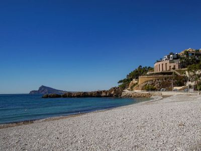 Playa de guijarros con aguas turquesas cristalinas y costa rocosa. Edificios modernos de fachadas reflectantes y una estructura redondeada bordean la ladera, bajo un cielo azul.