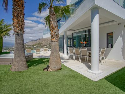 Terraza comedor exterior cubierta con mobiliario moderno, junto a un césped artificial y palmeras. La zona de jacuzzi y piscina ofrece vistas a la montaña y al mar.