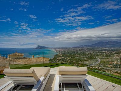 Terraza en la azotea con dos tumbonas blancas sobre tarima de madera clara y césped artificial, con vistas panorámicas al Mediterráneo, la costa de Altea y las montañas lejanas.