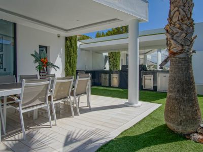 Terraza exterior con comedor al aire libre sobre suelo de lamas claras y césped artificial. Incluye una cocina exterior cubierta con barbacoa y electrodomésticos de acero inoxidable, junto a una palmera.
