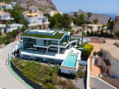 Vista aérea de una villa blanca moderna con piscina infinita, amplias fachadas de cristal, techo verde y jacuzzi, ofreciendo vistas a la montaña y al mar.