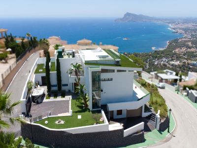 Vista aérea de una villa moderna blanca con arquitectura de varios niveles, techos planos verdes y ascensor exterior de cristal. La propiedad incluye entrada privada y ofrece vistas panorámicas al mar y la costa.
