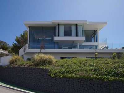 Vista exterior de villa moderna de tres niveles con amplias fachadas de cristal y barandillas de vidrio, elevada sobre un muro de piedra y vegetación junto a una carretera curva.