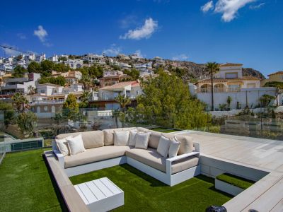 Zona de salón en la azotea con sofá en forma de L sobre césped artificial, mesa de centro blanca y barandillas de cristal. Vistas despejadas a la ladera escalonada con viviendas residenciales.