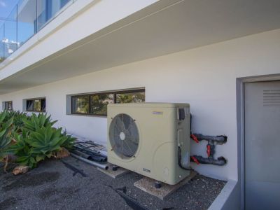 Zona técnica exterior de la villa con bomba de calor inverter AstralPool, ventanas horizontales y puerta de servicio. Agaves adornan la moderna fachada blanca.