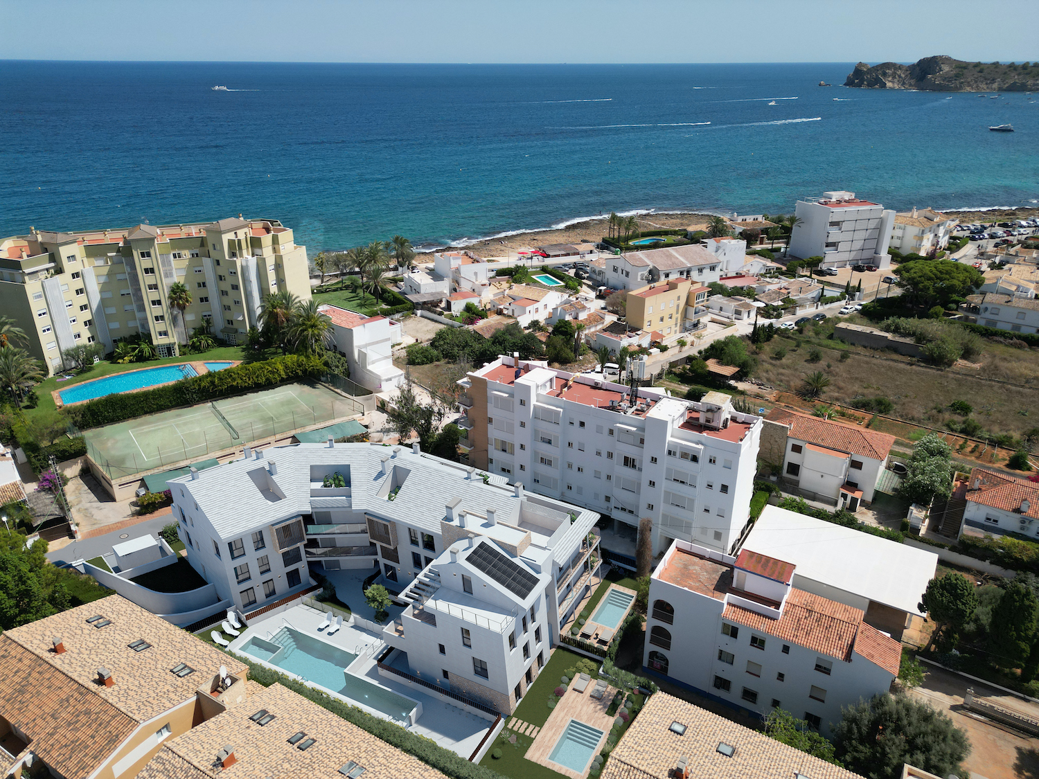 Vista aérea de la costa de Jávea, mostrando complejos residenciales con piscinas y pista de tenis, frente al Mediterráneo y un cabo lejano.