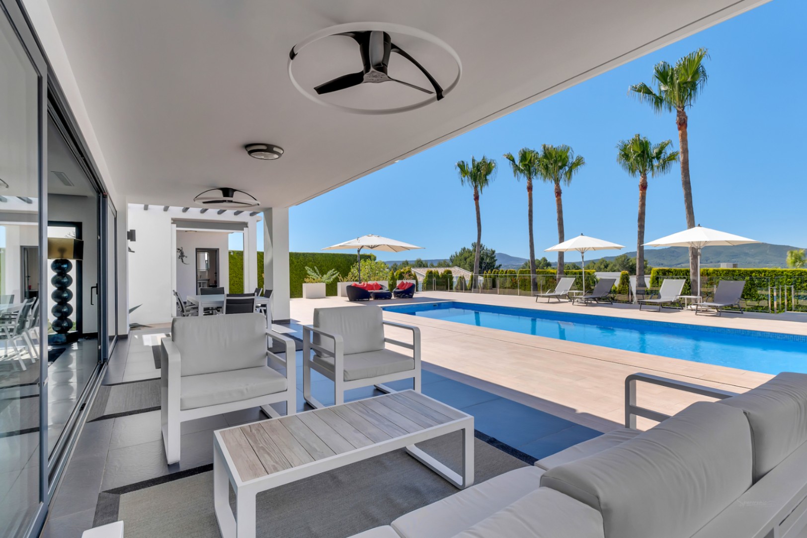 Amplia terraza cubierta con mobiliario lounge blanco, ventiladores de techo y puertas correderas, conectando directamente con la piscina y vistas a las montañas.