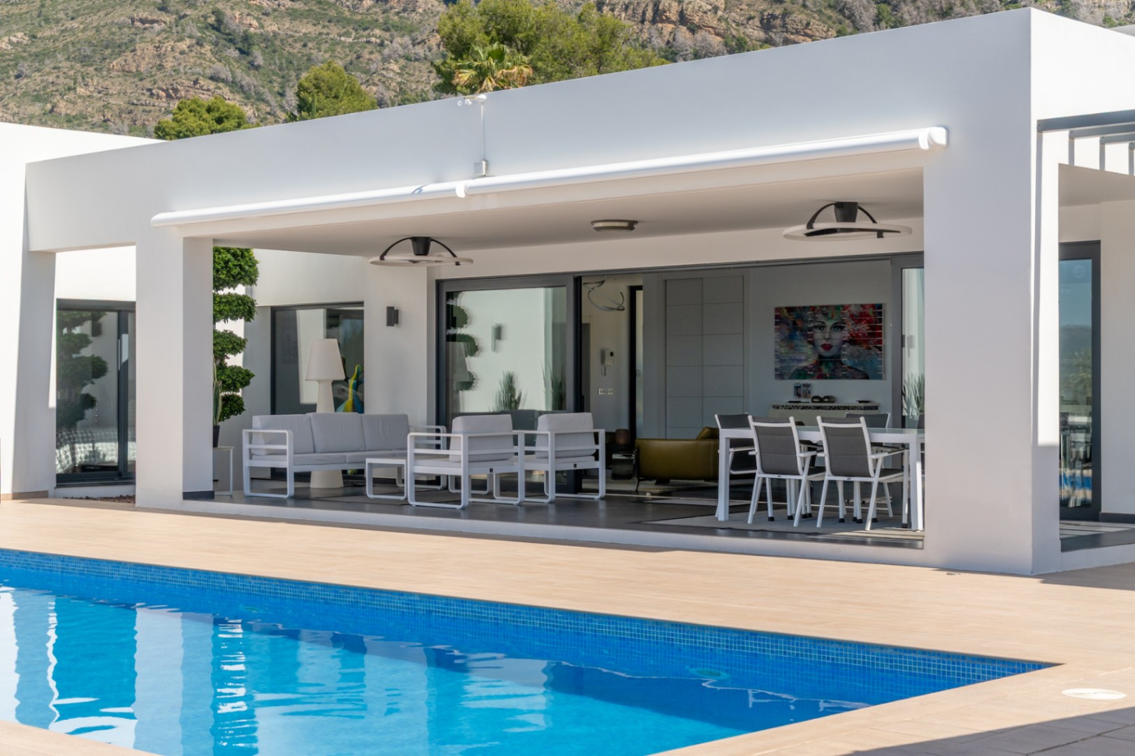 Amplia terraza cubierta con toldo retráctil, mobiliario de exterior y comedor, conectada a la piscina y al interior con vistas a la montaña.