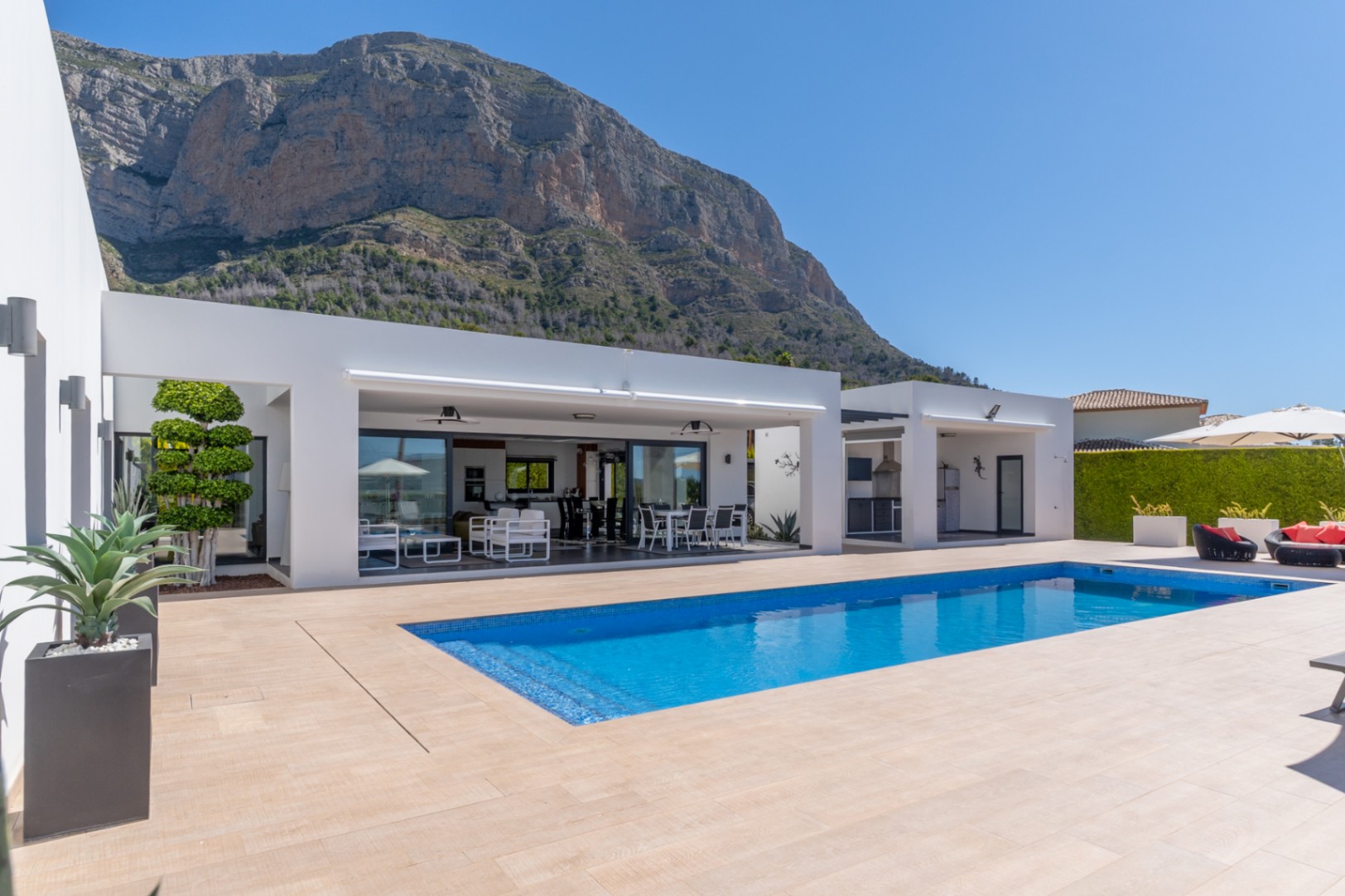 Amplia zona exterior con piscina rectangular de mosaico azul, terraza de baldosas claras y villa moderna blanca con salón y comedor cubiertos integrados, con el Montgó de fondo.