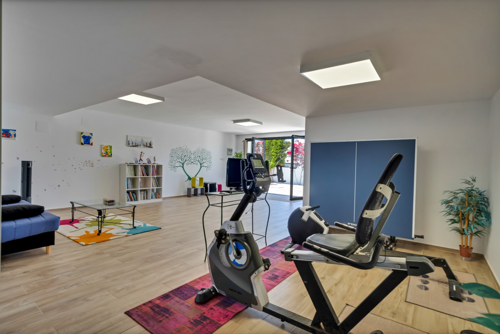 Amplio espacio inferior con equipamiento de gimnasio, una acogedora zona de estar con sofá azul y estantería. Acceso directo al exterior mediante puertas de cristal y suelo de madera clara.