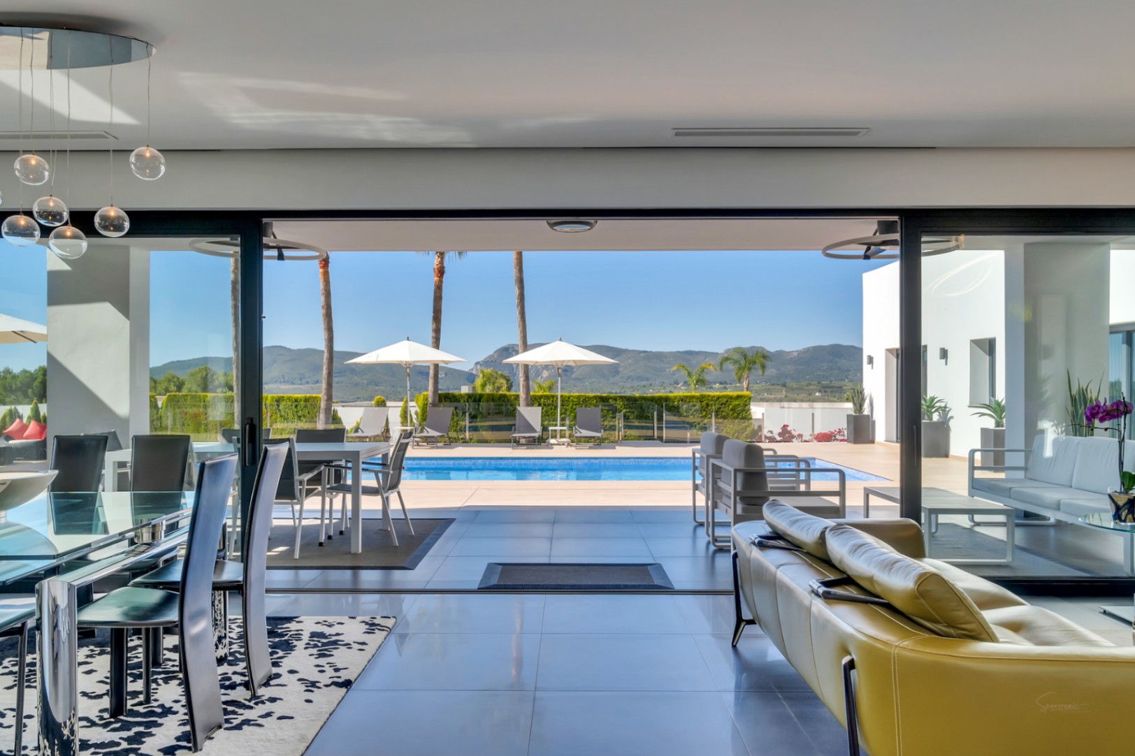 Amplio salón-comedor con mesa de cristal, lámparas colgantes modernas y sofá amarillo, que se abre a la terraza de la piscina con tumbonas y vistas a las montañas lejanas.
