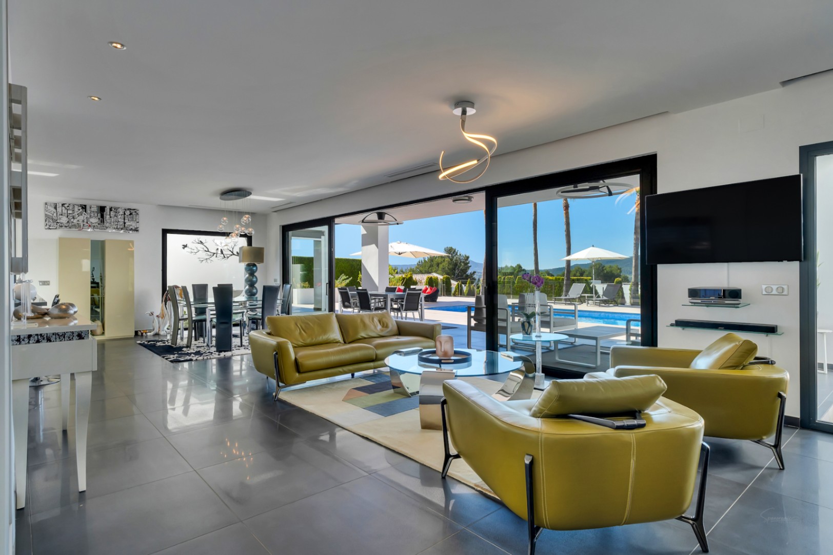 Amplio salón-comedor de planta abierta con acceso directo a la terraza y piscina climatizada mediante grandes puertas correderas. Mobiliairo verde oliva y suelo de gres oscuro.