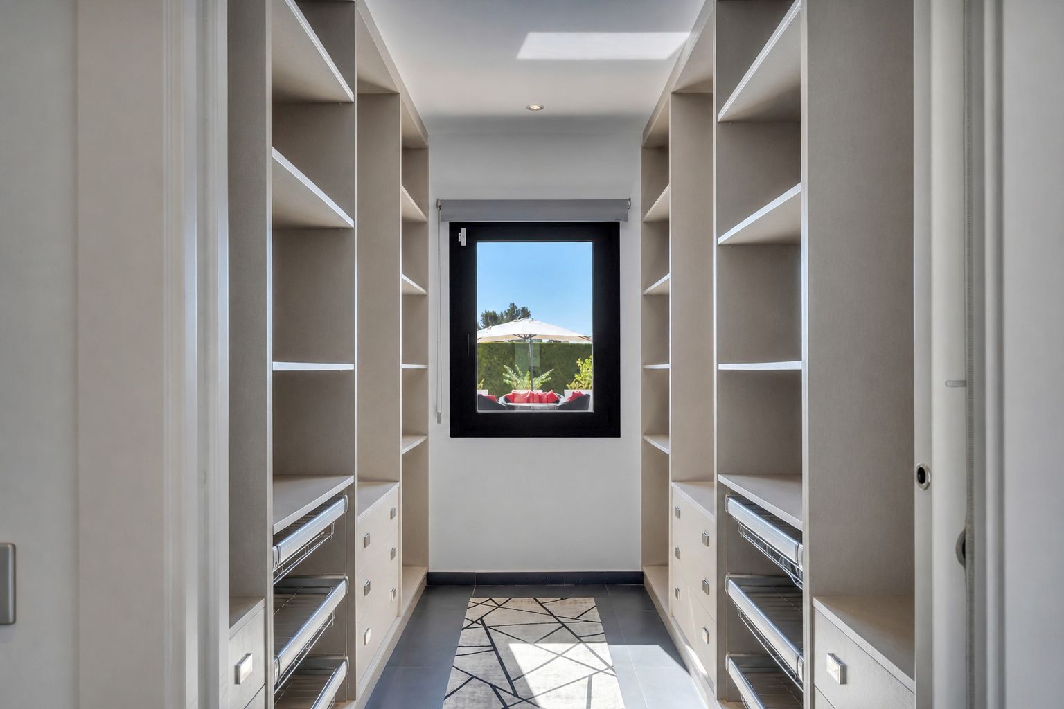 Amplio vestidor con estanterías integradas, cajones y cestas extraíbles, iluminado por luz natural de una ventana con vistas al jardín privado.