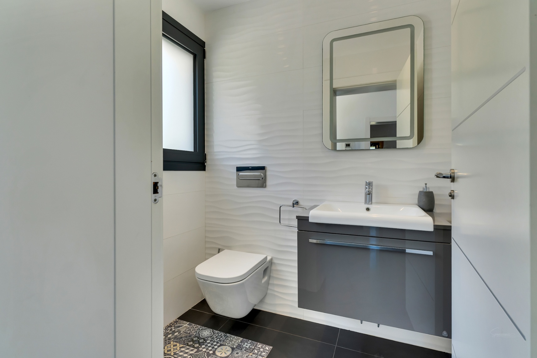 Aseo moderno con inodoro suspendido, mueble de lavabo flotante gris y azulejos blancos texturizados. Destacan el espejo retroiluminado y el detalle de suelo hidráulico.