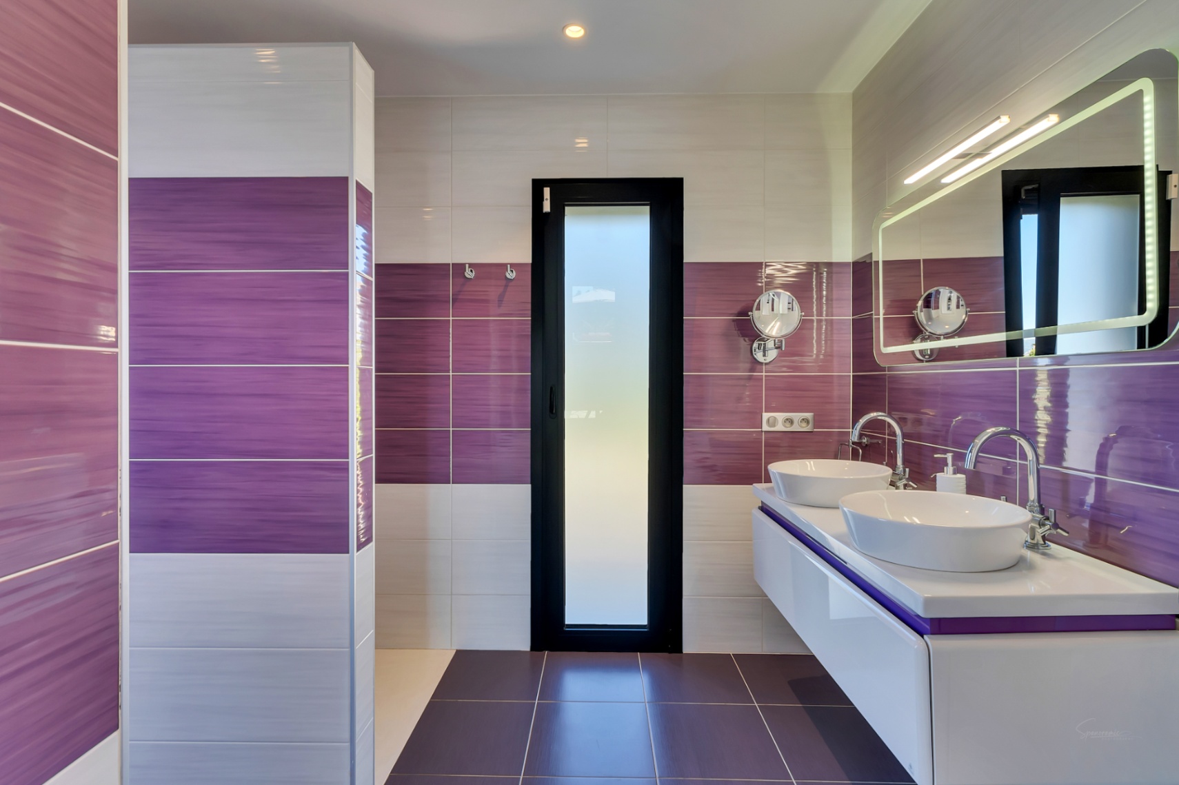 Baño contemporáneo con azulejos morados y blancos, tocador doble flotante con lavabos ovalados y espejo iluminado. Una puerta de cristal esmerilado aporta privacidad.