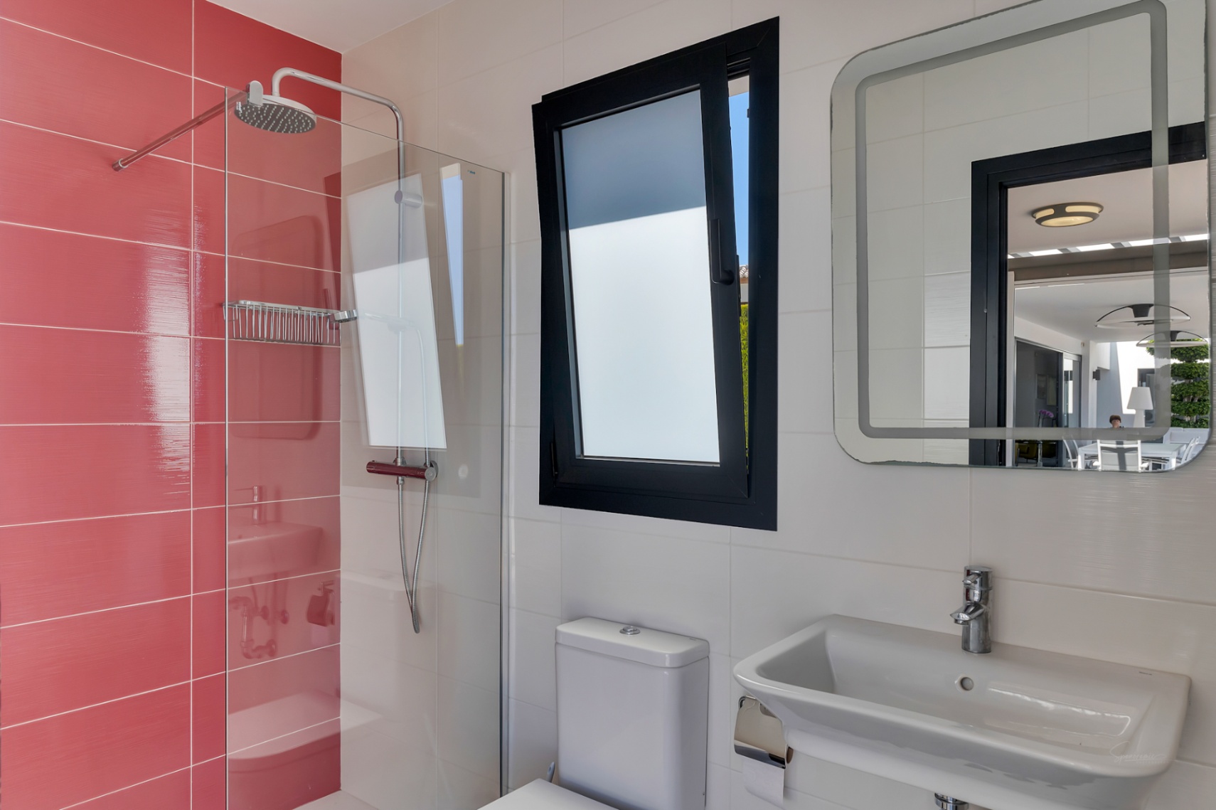 Baño funcional con ducha de plato de lluvia y azulejos rojos, ventana oscilobatiente y espejo iluminado que refleja la terraza exterior.