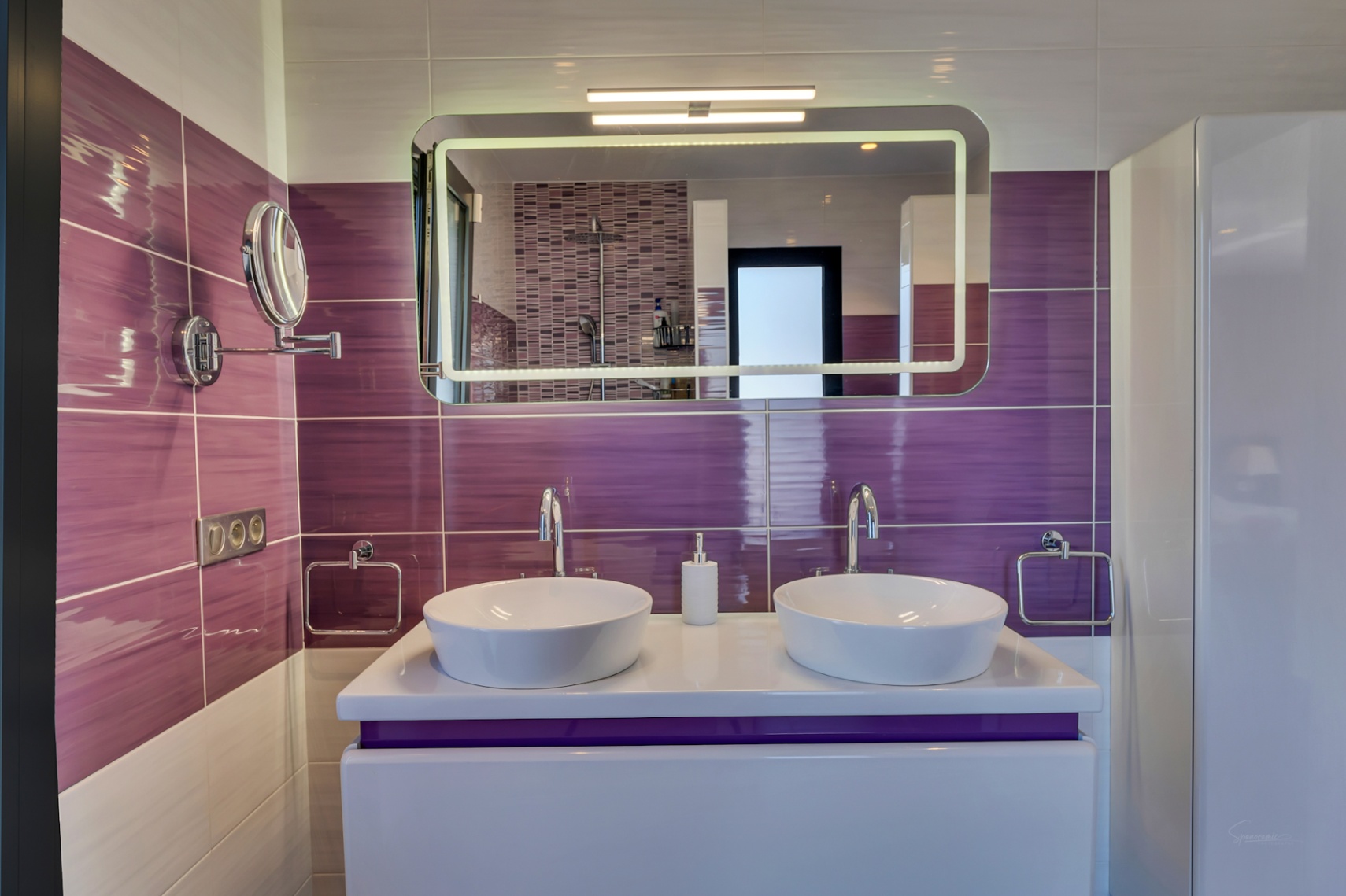 Baño moderno con doble lavabo sobre encimera, azulejos morados horizontales y un espejo iluminado que refleja la ducha con mosaico.