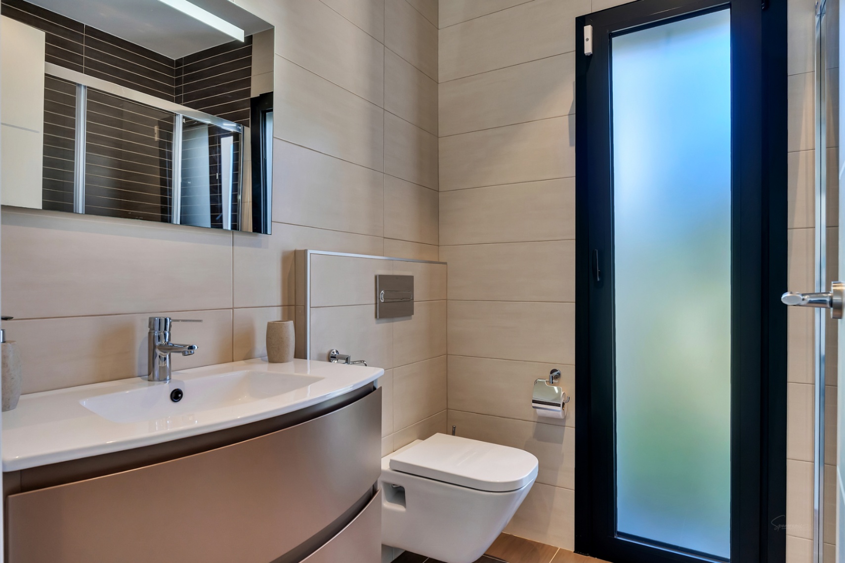 Baño moderno con inodoro suspendido, lavabo con mueble bajo y grifería cromada. Paredes alicatadas en tono neutro y puerta con cristal esmerilado para luz natural.