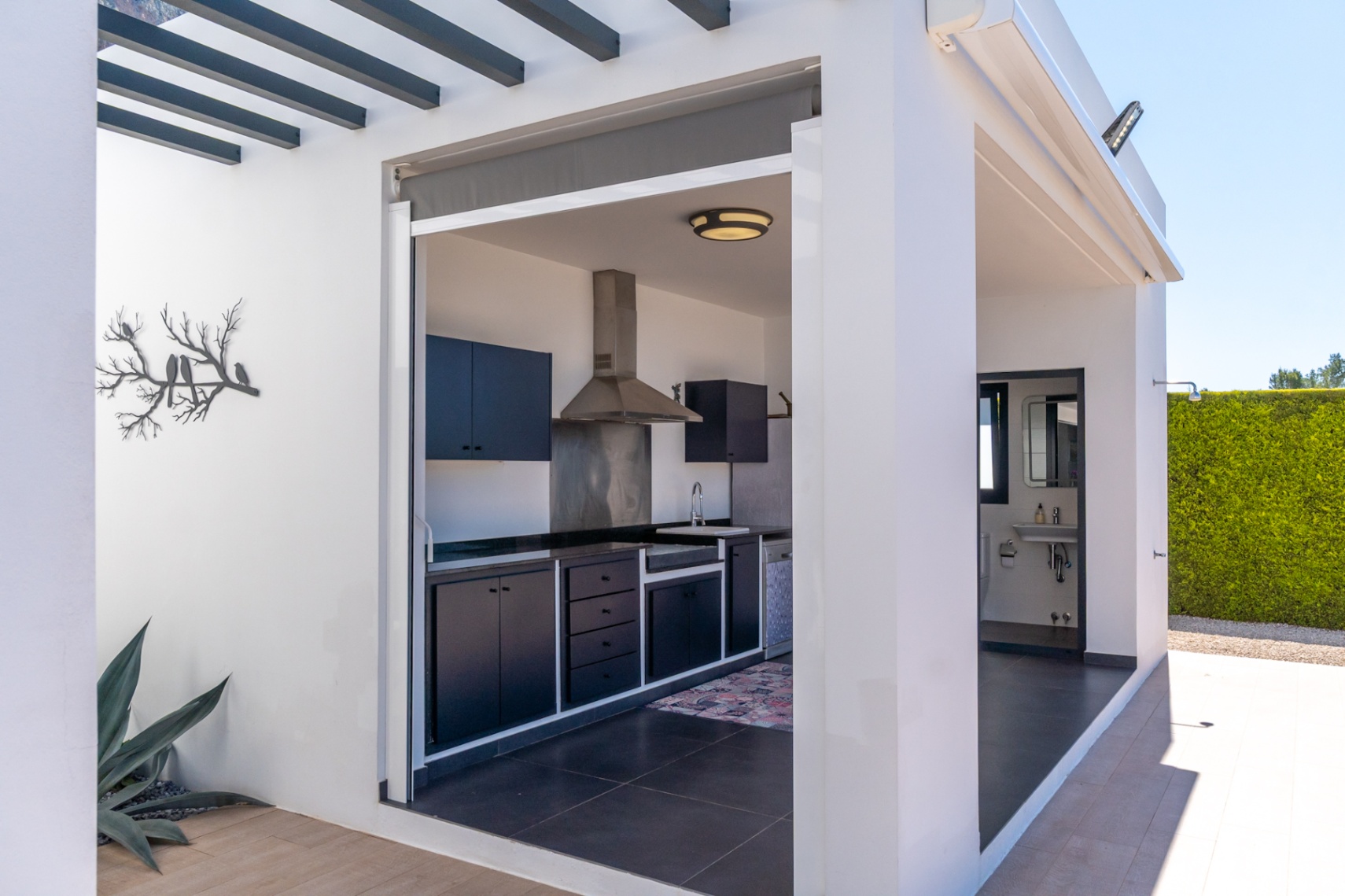 Cocina exterior con gabinetes oscuros y encimeras, equipada con extractor de acero y fregadero, adyacente a un práctico baño exterior y un seto de privacidad.