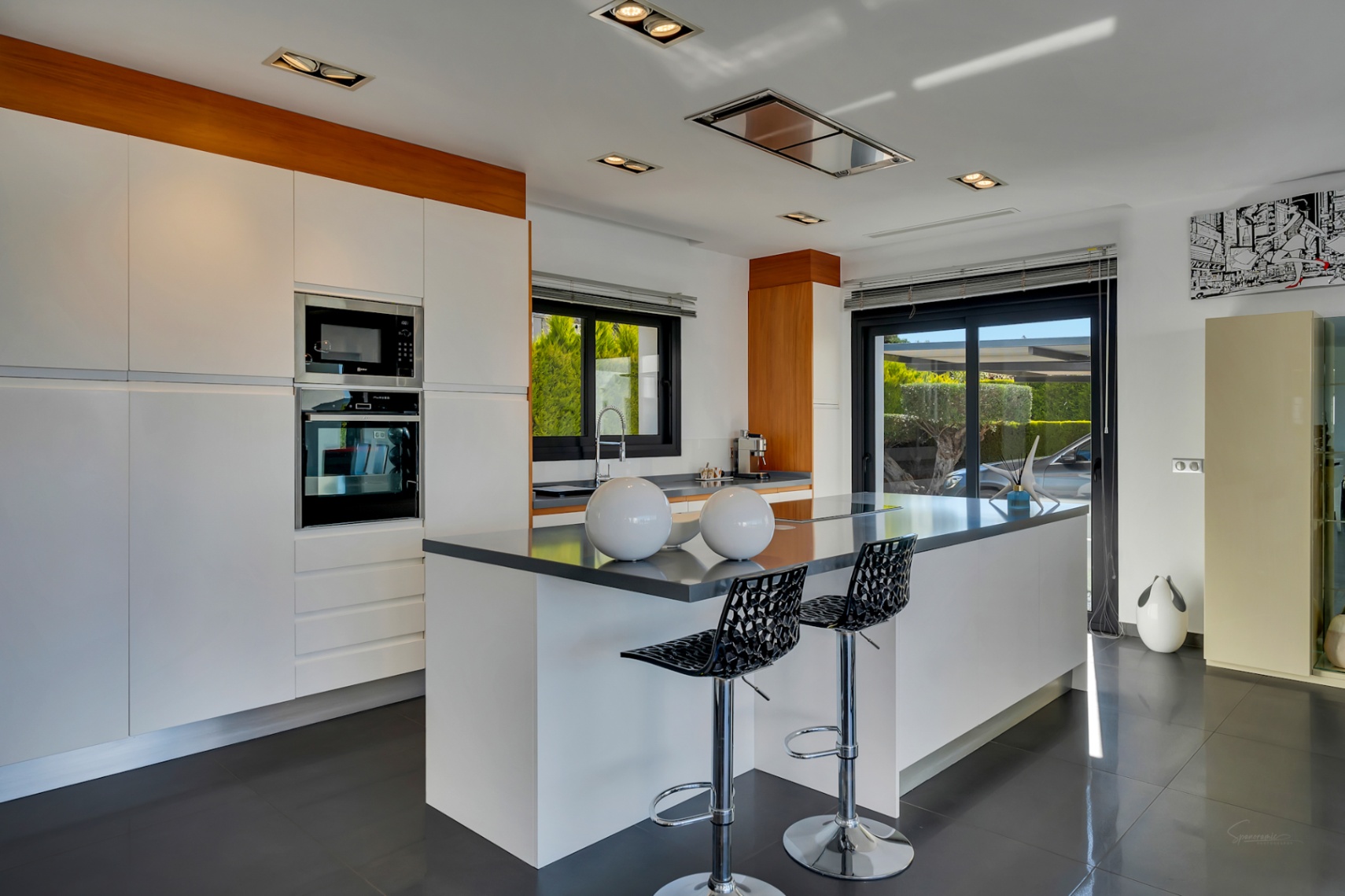 Cocina moderna con gran isla central y asientos, electrodomésticos integrados, armarios blancos y acceso directo al exterior mediante puerta corredera de cristal.
