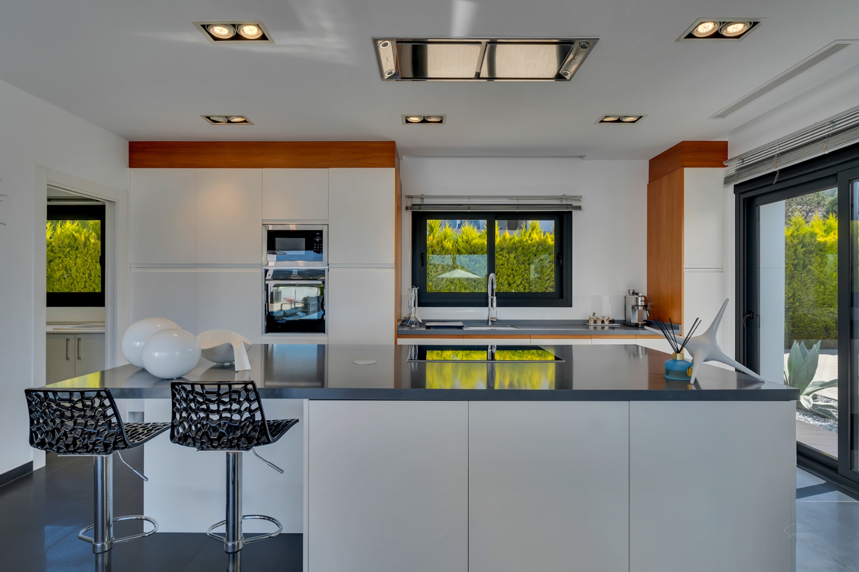 Cocina moderna con mobiliario blanco y encimeras oscuras, destacando una isla central con asientos. Electrodomésticos integrados y luz natural se combinan con vistas a la vegetación exterior.