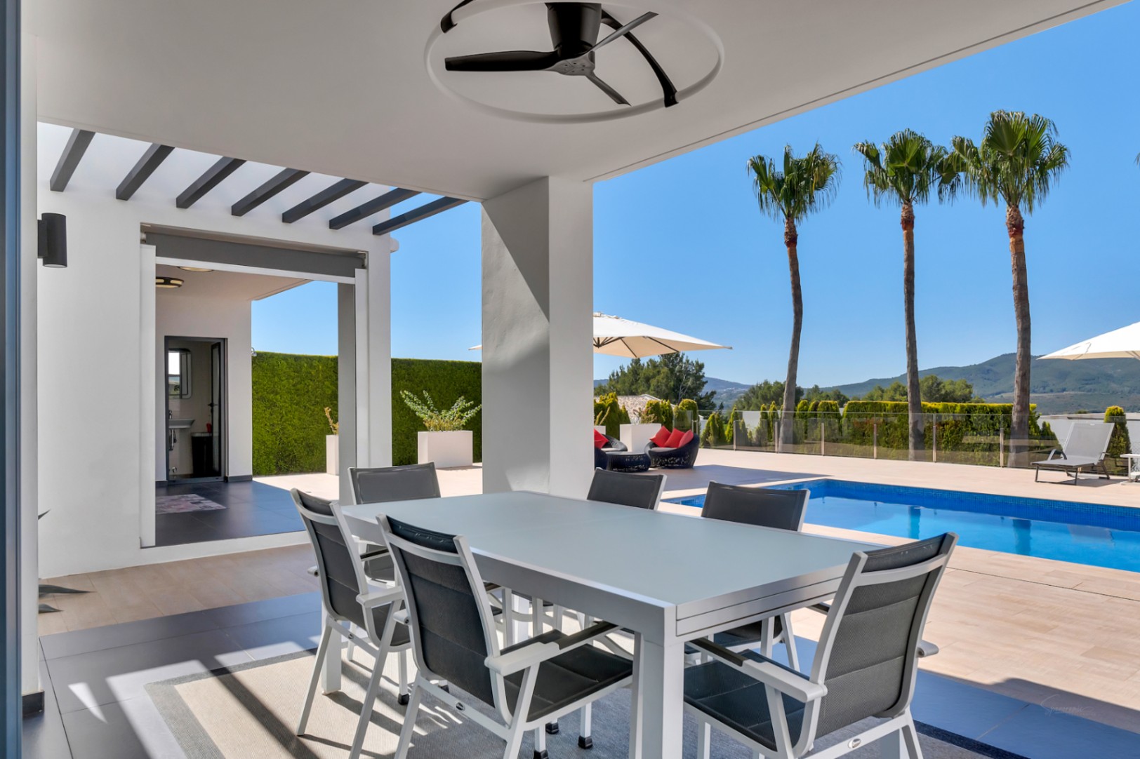 Comedor exterior cubierto con mesa y sillas modernas, ventilador de techo, vistas a la piscina, palmeras y montañas, con un baño exterior adyacente.