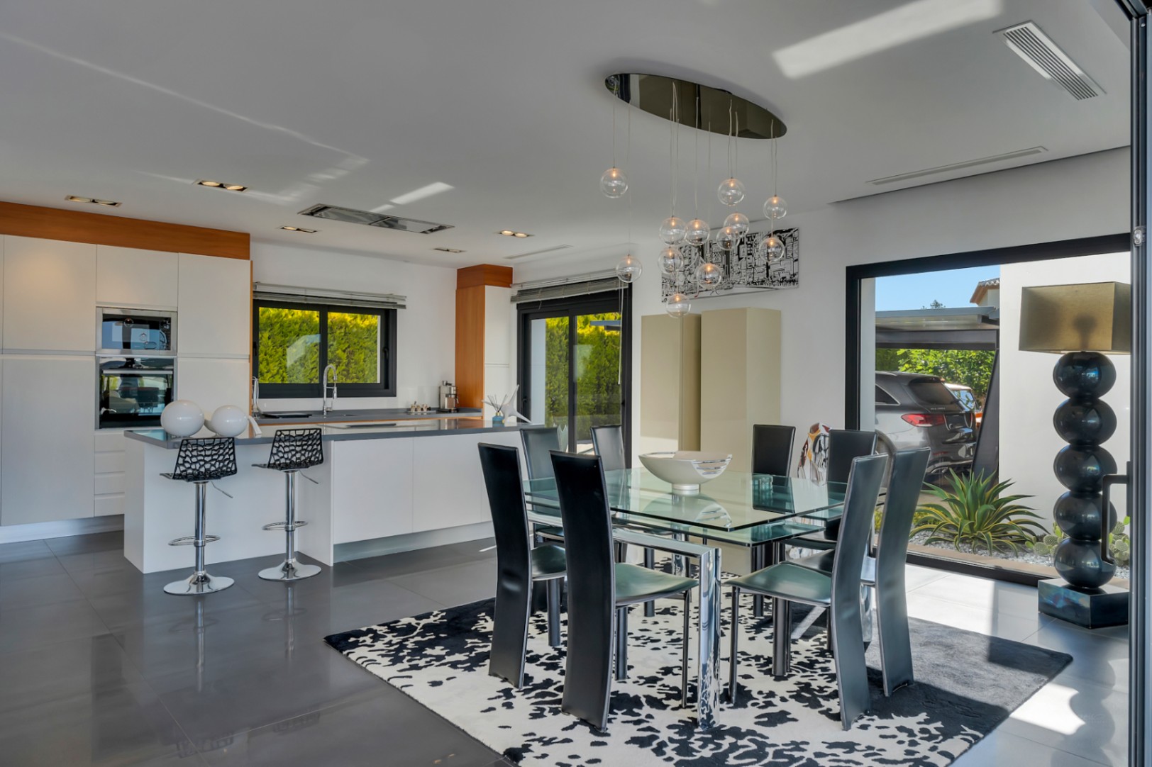 Comedor integrado con cocina moderna de diseño, mesa de cristal y sillas negras, bajo una lámpara colgante contemporánea. Grandes puertas correderas conectan con el exterior.