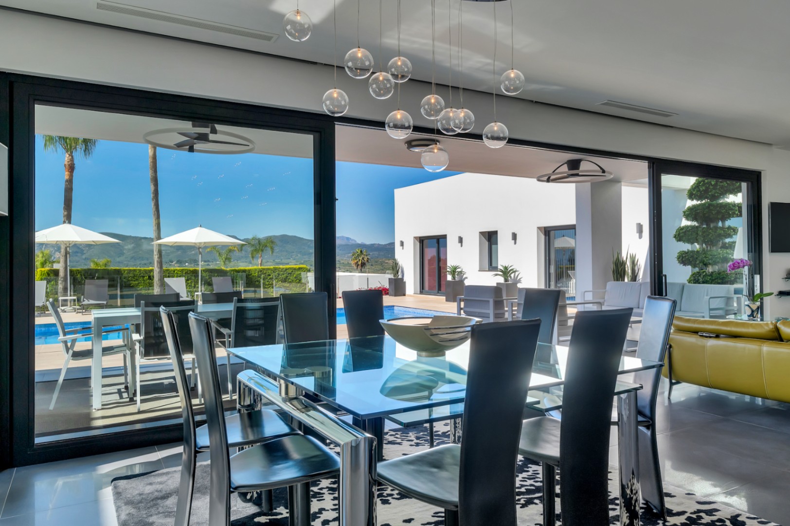 Comedor moderno con mesa de cristal y sillas negras, conectado a una terraza con piscina, tumbonas, palmeras y vistas a la montaña.