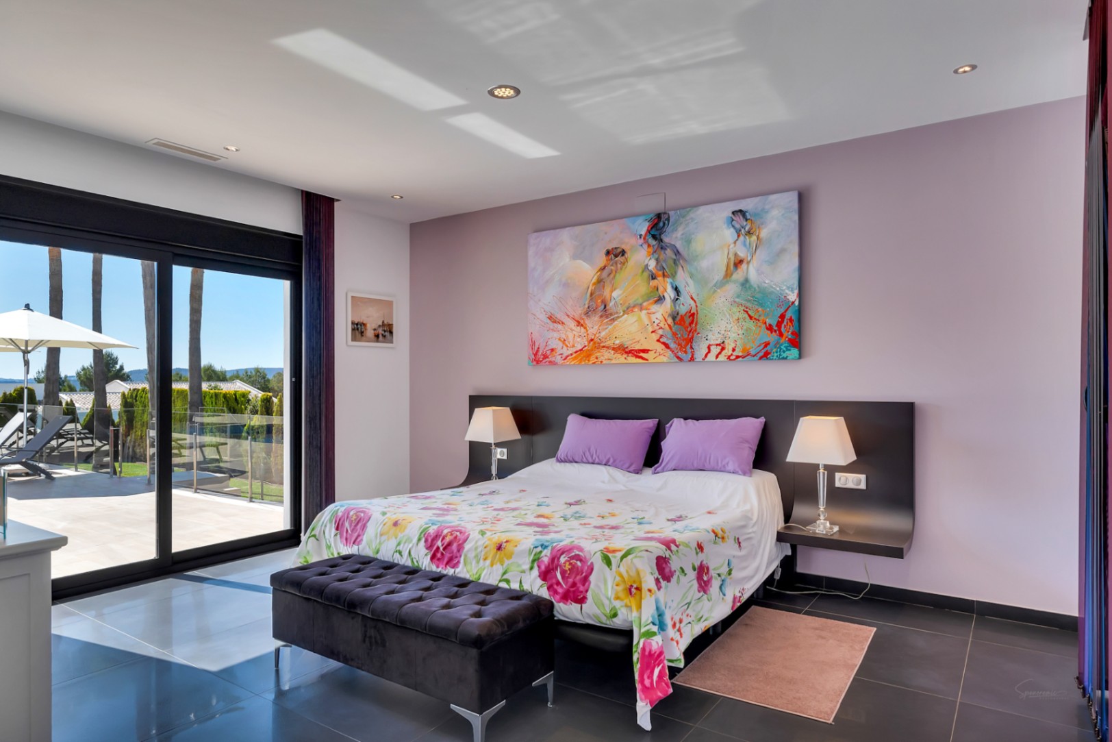 Dormitorio con acceso directo a la terraza a través de grandes puertas correderas de cristal, suelo de baldosas oscuras, edredón floral y una colorida obra de arte abstracta. Vistas al exterior con palmeras.