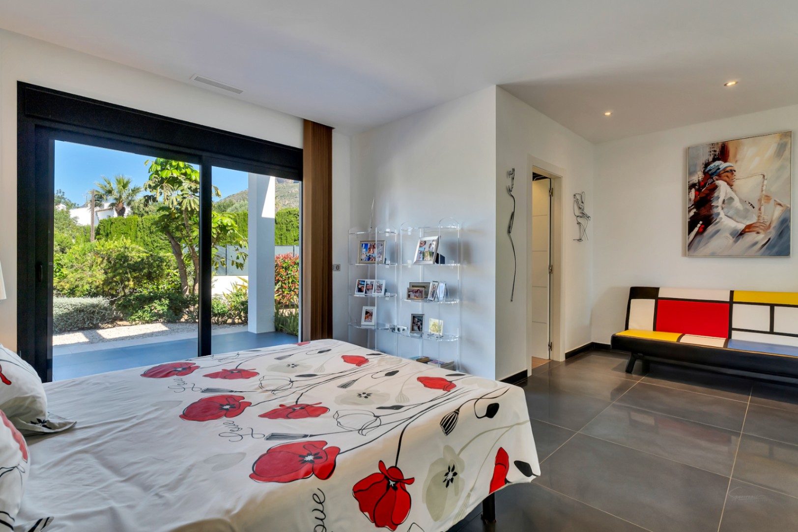Dormitorio con grandes puertas correderas de cristal que abren a un jardín mediterráneo exuberante, suelo de gres oscuro y mobiliario contemporáneo.