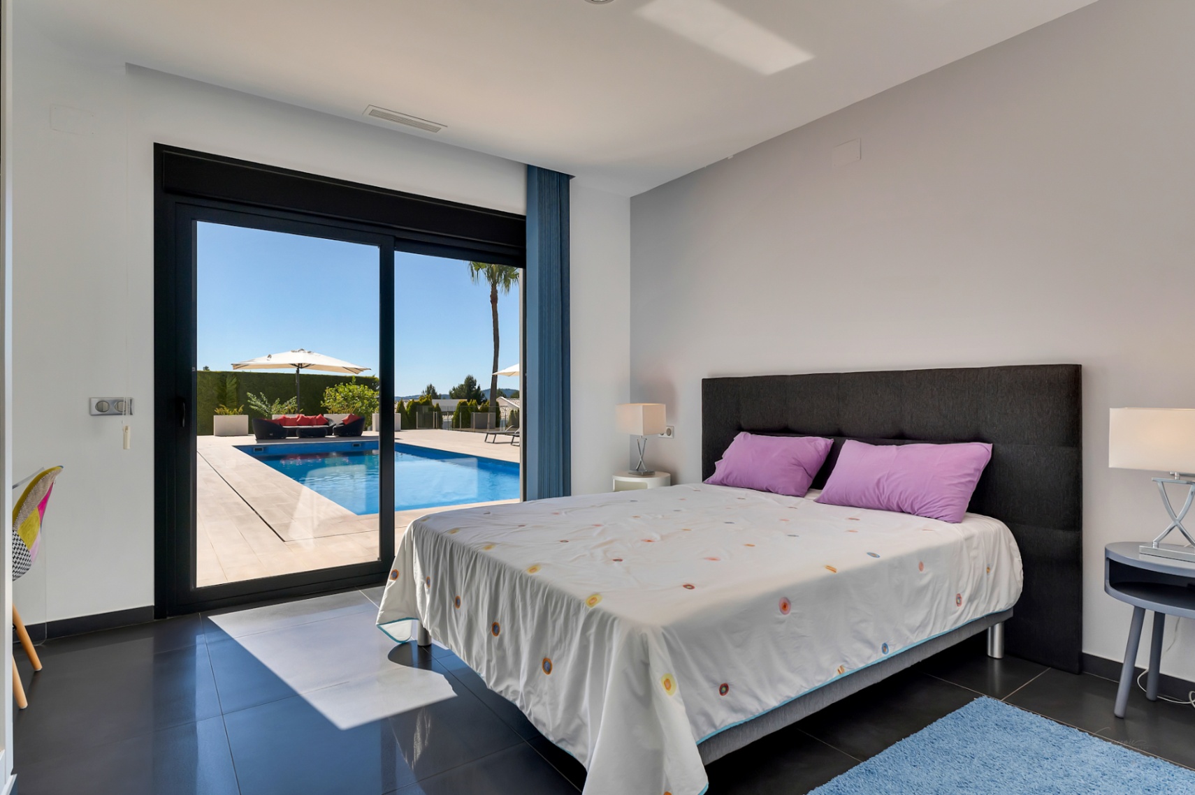 Dormitorio con suelos de baldosas oscuras y acceso directo a la piscina, que incluye una zona de relax exterior con sofá rojo y sombrilla bajo un cielo despejado.